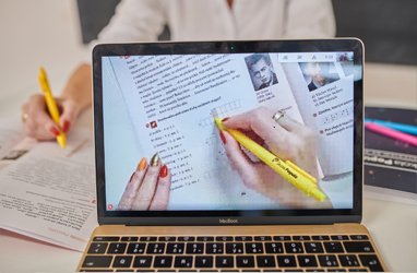 Pedagog ukazuje cvičení v učebnici kterou snímá telefon. Kamera z notebooku míří na něj.